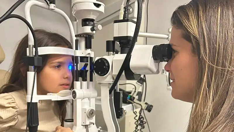 Criança apoiada na lâmpada de fenda durante exame oftalmológico, com a médica avaliando os olhos do outro lado do aparelho.